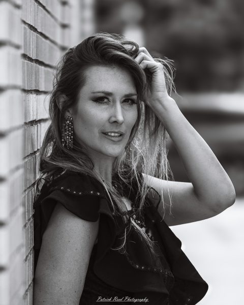 Black-and-white portrait of a woman leaning against a brick wall with one hand in her hair.